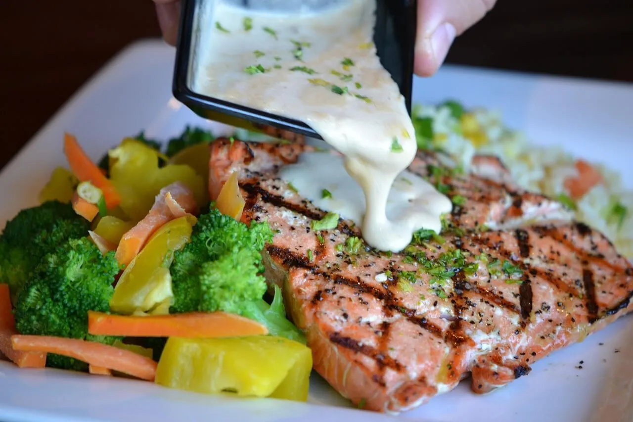 Hand pouring white sauce over a grilled salmon steak with vegetables on a white plate
