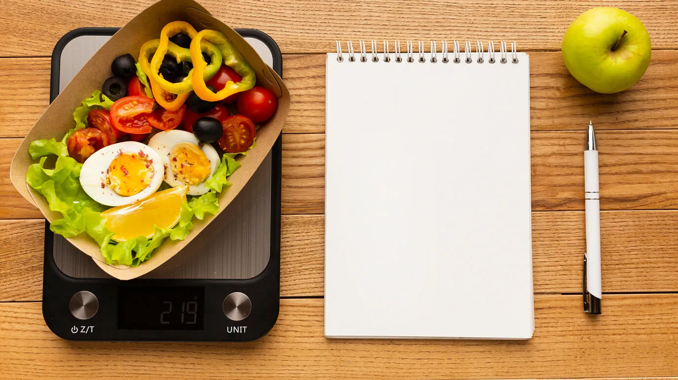 Kitchen scale with a portion of food and a notebook used to calculate daily calories